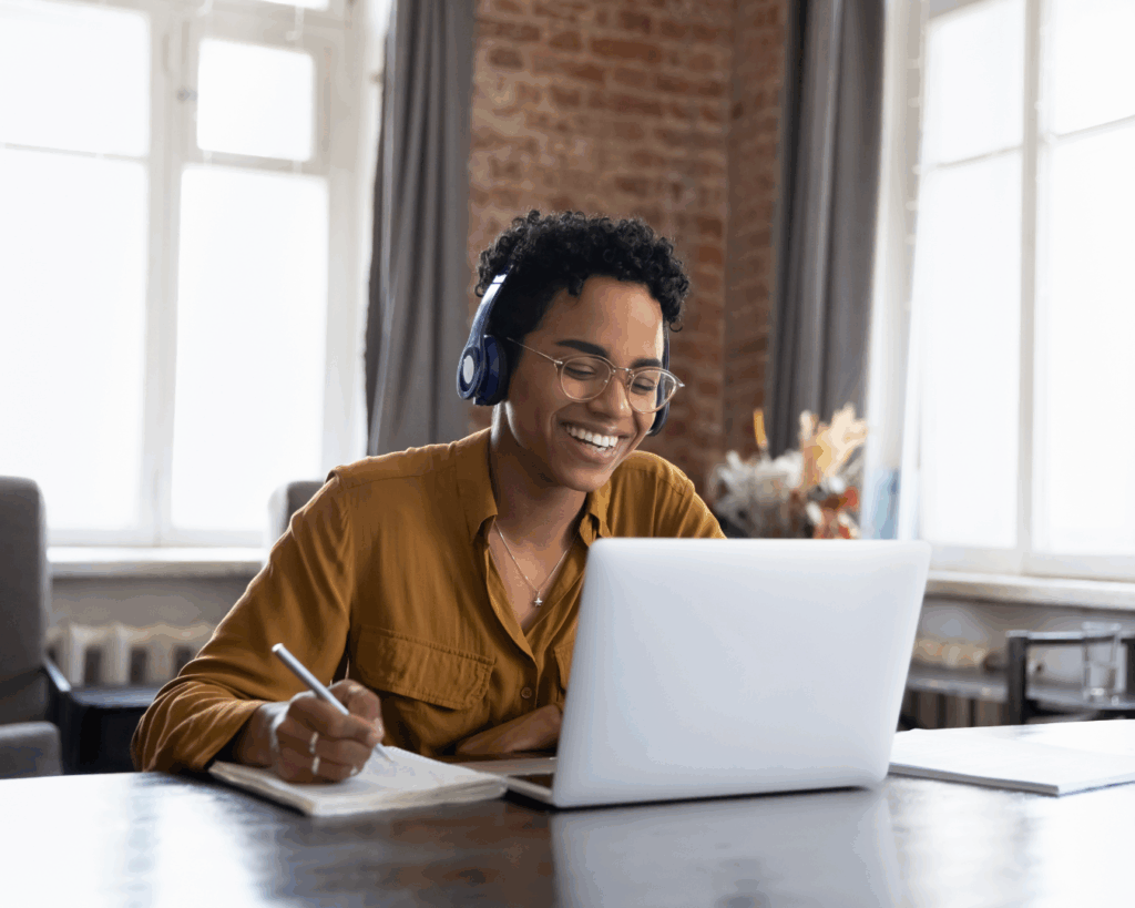 Cheerful woman working on a laptop, wearing headphones and writing on a notepad