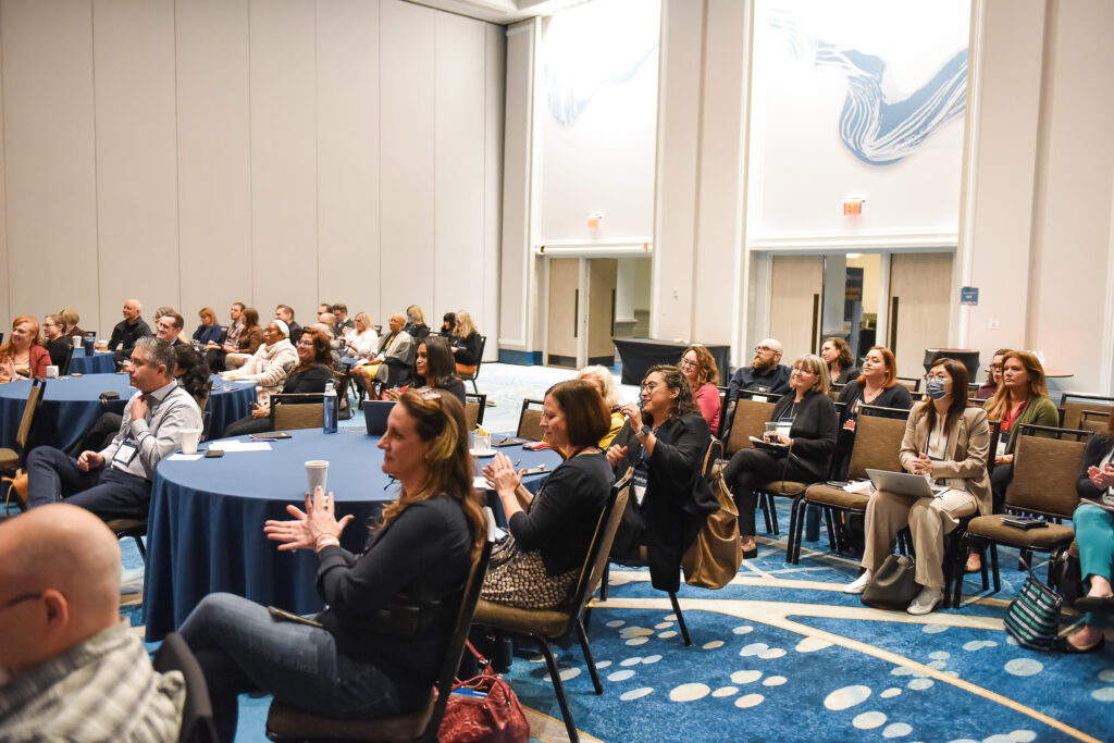 Photo of the interior of a crowded conference room, with seated attendees facing the front of the room, clapping.