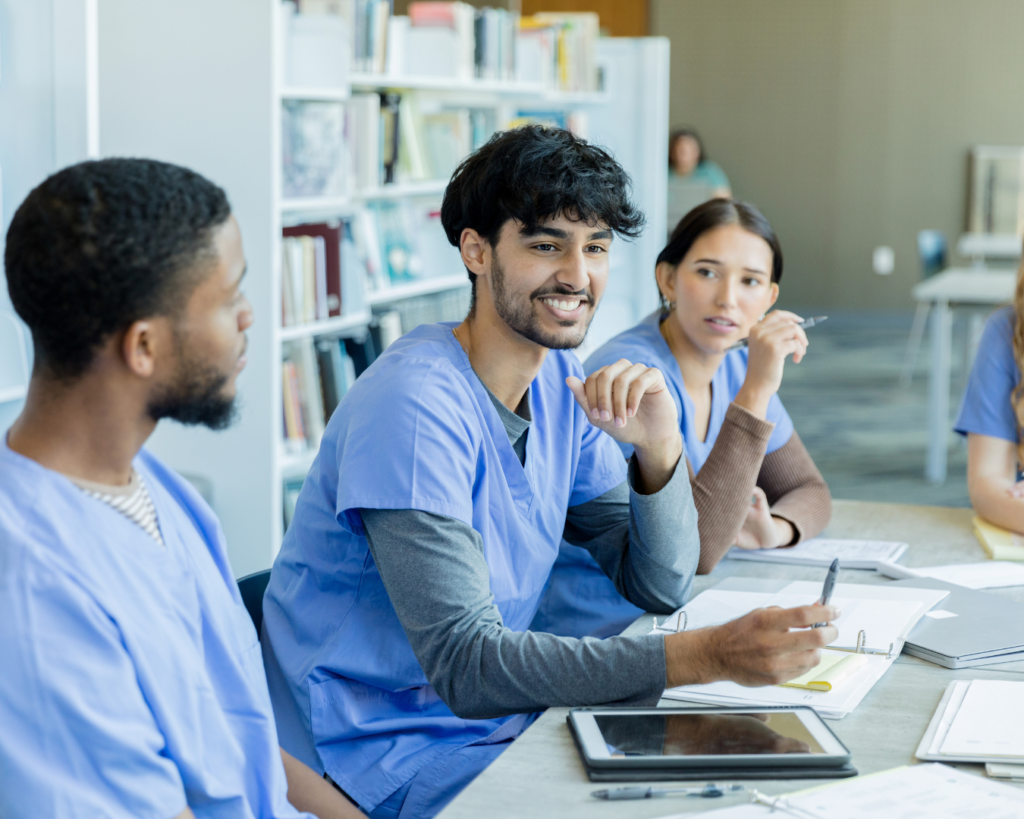 Medical students in scrubs sit around a study table, discussing happily.