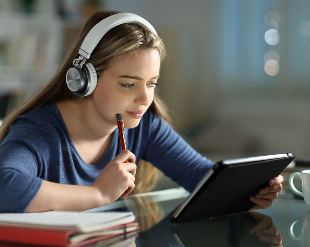 A young college student studies on her tablet, wearing headphones, and deep in thought.