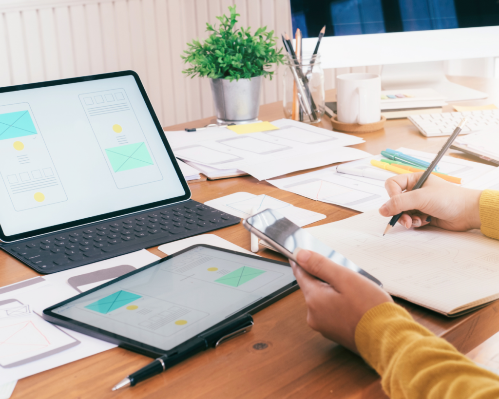 The hands of a visual designer as they work at a messy desk top. Papers with wireframes are scattered around a laptop, and the designer sketches on a pad, referencing their smartphone and tablet.