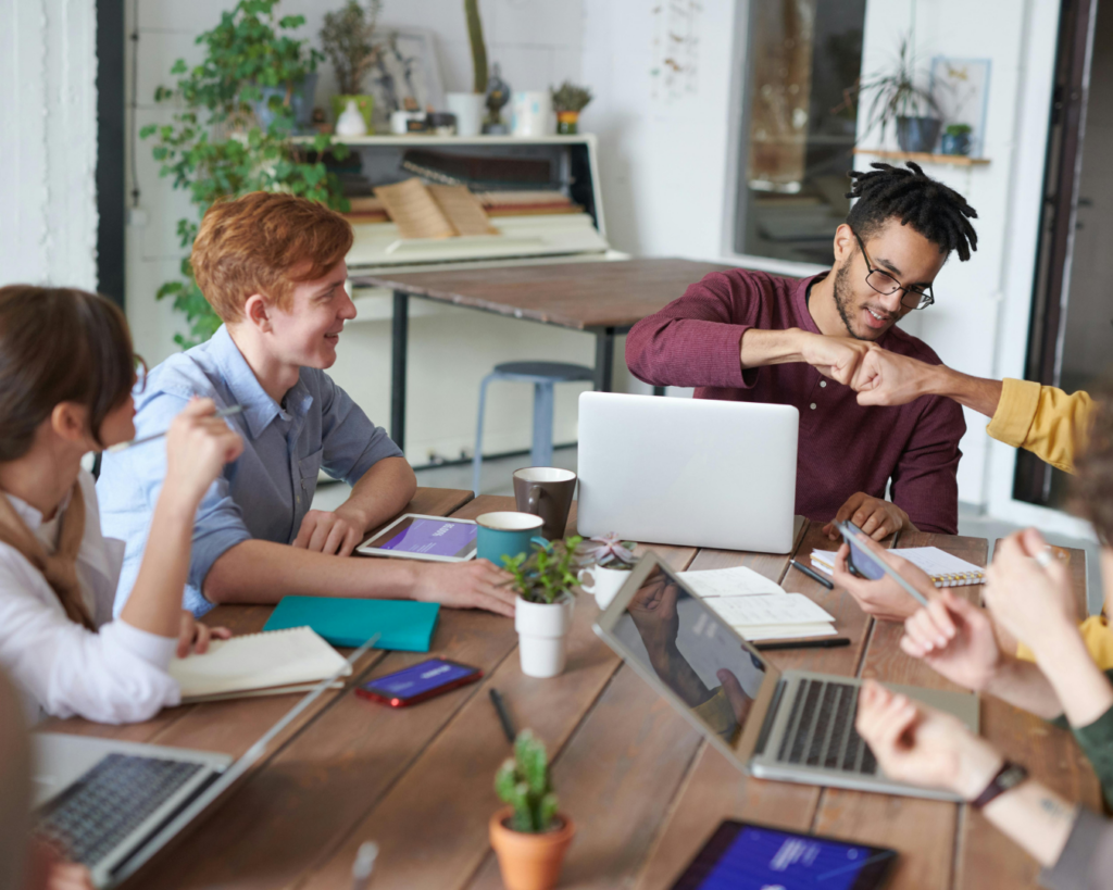 A small, diverse team of professionals collaborate around a table, working on laptops. Two team members share a fist bump.