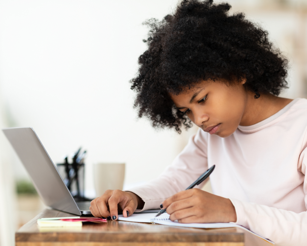 A young girl is doing homework at a desk. Sitting in front of an open laptop, she is intently writing on a piece of paper.