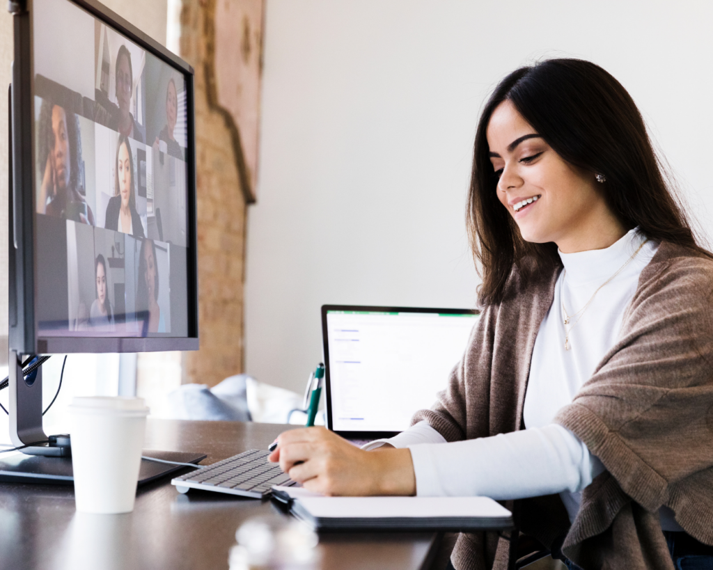 A woman takes a video-call meeting from her desk, writing in a notebook, her coworkers are displayed in a gallery on her desktop monitor.