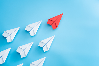 Several white paper planes in an arrow-formation, being led by a single red paper plane.