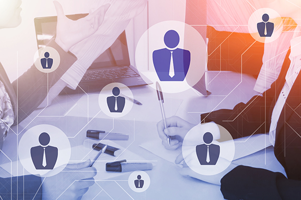 The hands of several people working at a shared meeting table, overlaid with icons of a businessperson and interconnected lines.
