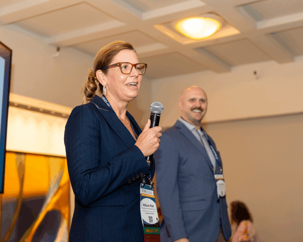 Photo of Allison Rief and Cody House speaking on a stage at the Accelerate 2025 closing ceremony. Allison speaks into a microphone, Cody smiles, standing next to her.