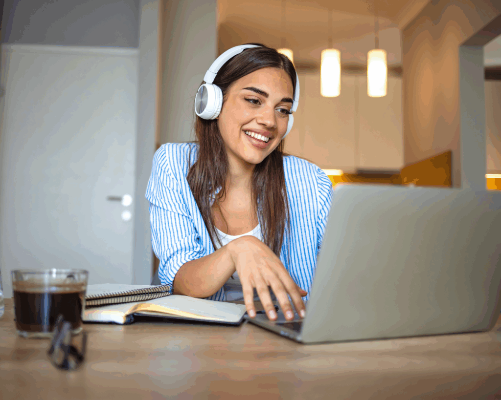 A photo of a female student wearing headphones, smiling and talking towards her laptop.
