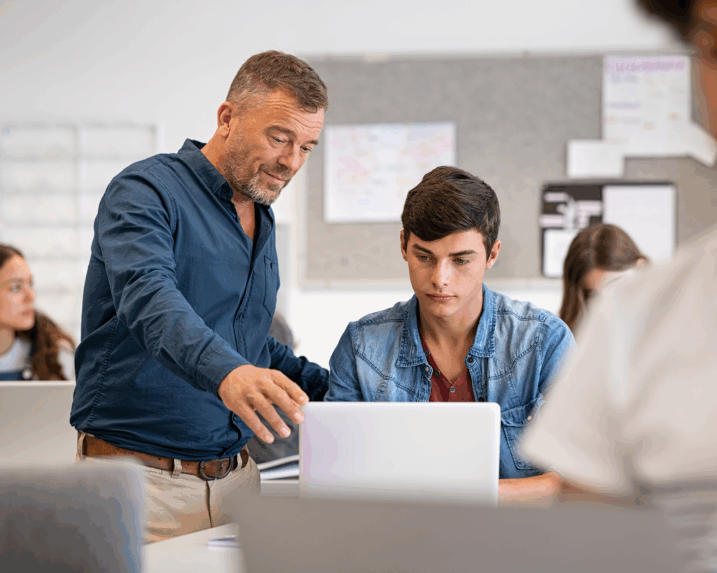 A male teacher gestures to a college-age student's laptop screen as he watches attentively.