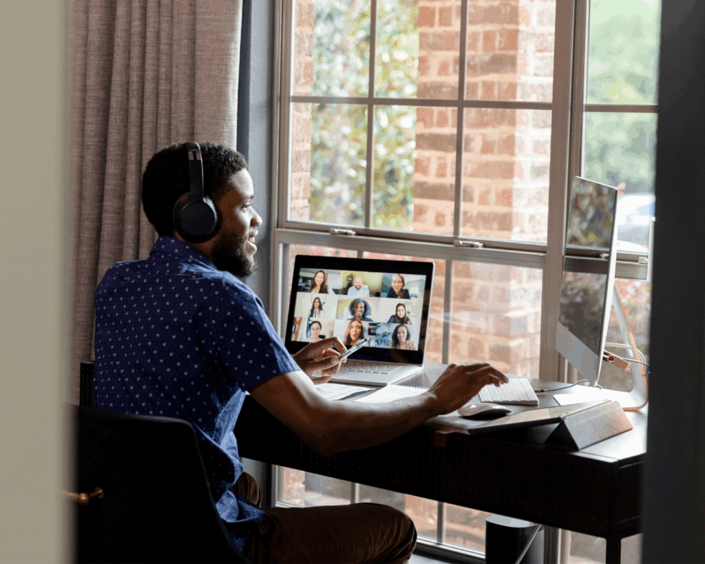 A man in a home office sits at a desk in front of a bright window. He wears headphones, and his laptop screen displays a gallery of virtual meeting attendants. He smiles, speaking in the meeting while hovering his hand over a keyboard attached to an additional computer monitor.