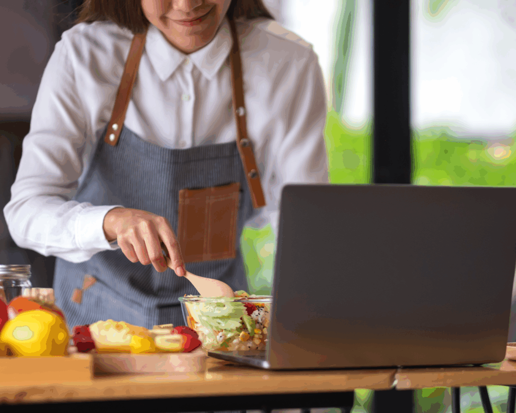 A woman in an apron mixes a bowl of vegetables on a table top in front of an open laptop.