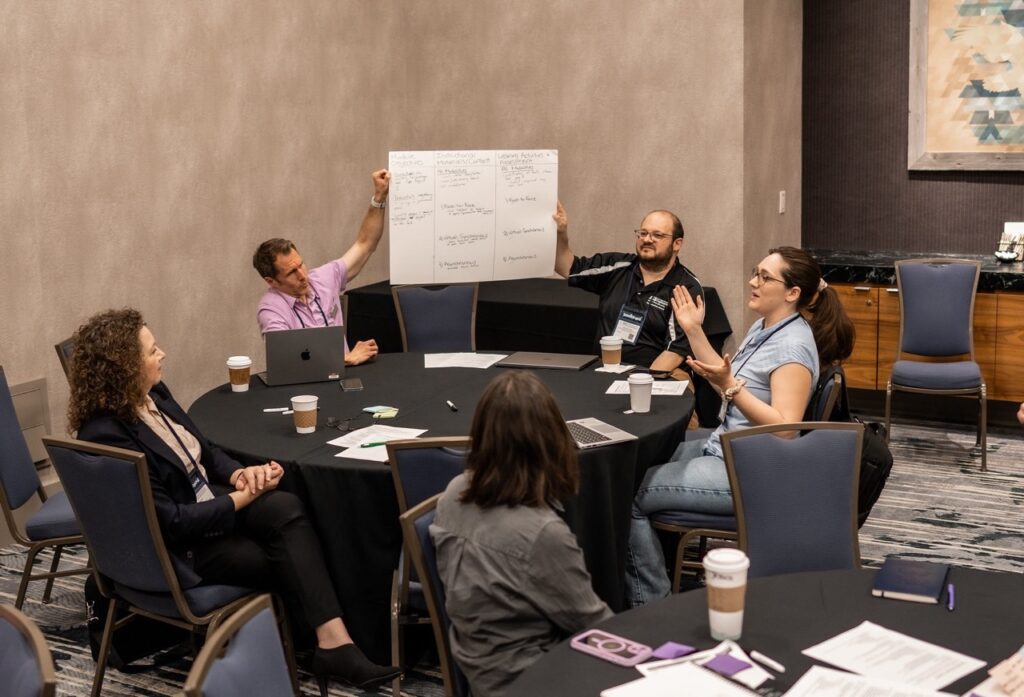Photo of a group of conference attendees at a round table, two hold up a poster-board with a marker-drawn diagram, a woman at the table talks, gesturing with her hands, and two other participants watch
