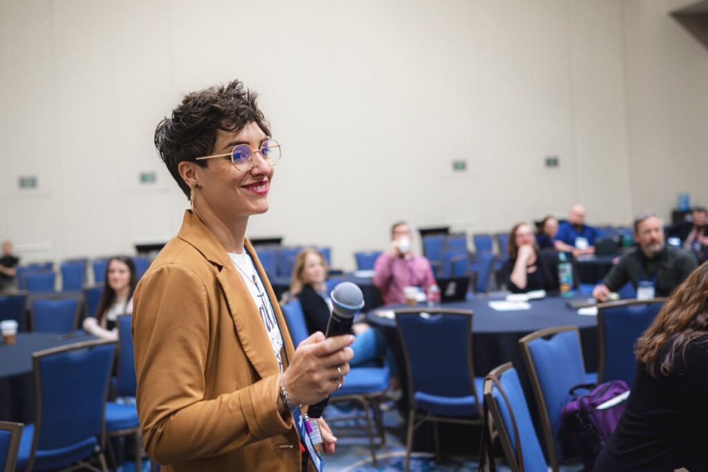 A photo of a woman presenting at an OLC event to a filled conference room, she's smiling, wearing a casual taupe blazer, and holding a microphone