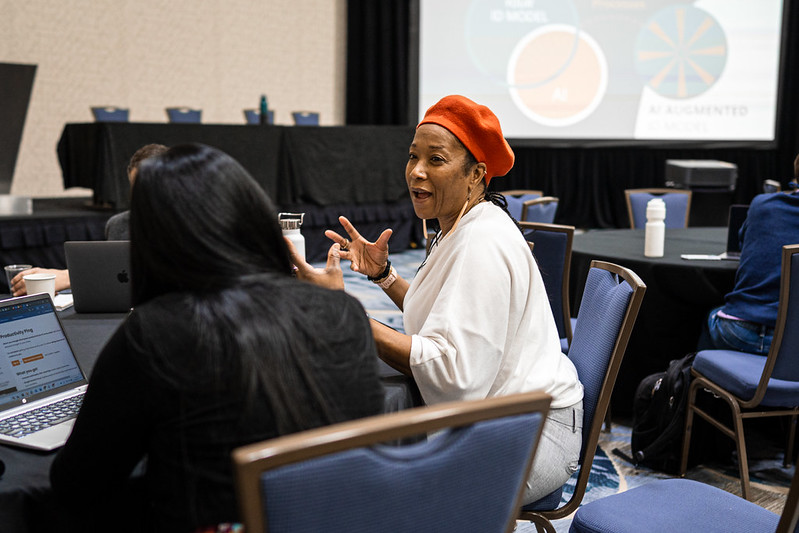 Photo of a woman in a head wrap sitting at a table at an OLC event, she speaks to another woman also seated, smiling and gesturing with her hands