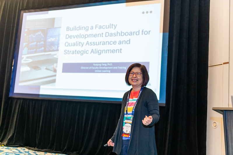 Photo of a presenter at OLC Accelerate standing in front of her presentation projected on to a large screen, gesturing towards it and smiling.