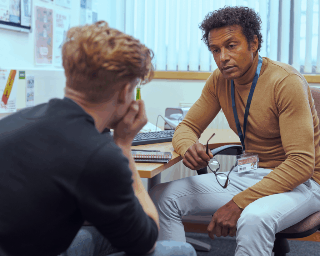 Photo of two men sitting in an office, one man leans toward the other with a stern expression, while the other rests his head in his hands.