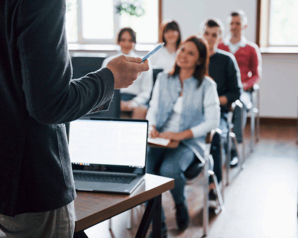 Photo from behind a man presenting at a short table with his laptop to a room of smiling adults.