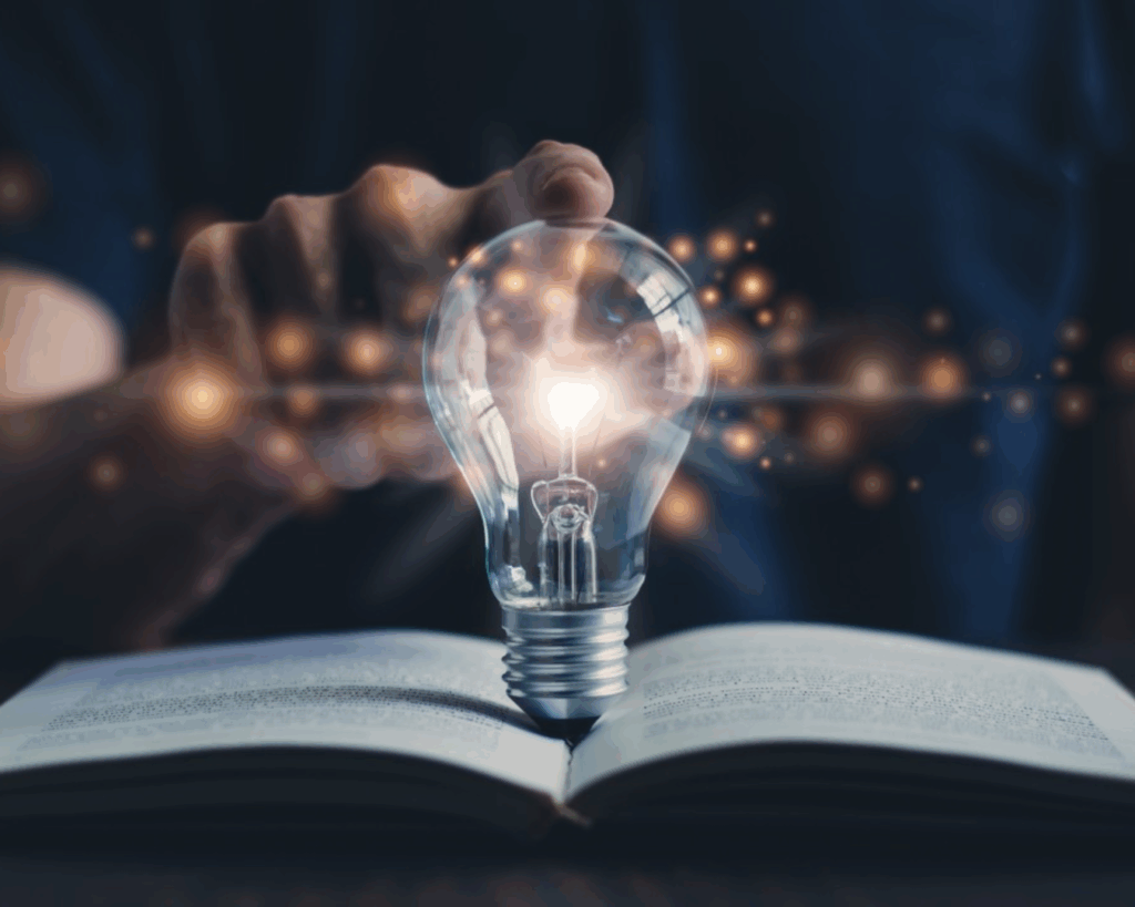 A dramatically lit image of a glowing lightbulb standing in the fold of an open book, with a person's hand reaching forward to lay a single finger on the top of the bulb.