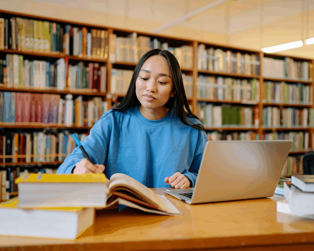 Image of a college-age girl working at a desk in a warmly-lit library. She has her laptop open and a stack of books in front of her, and she writes in a book.