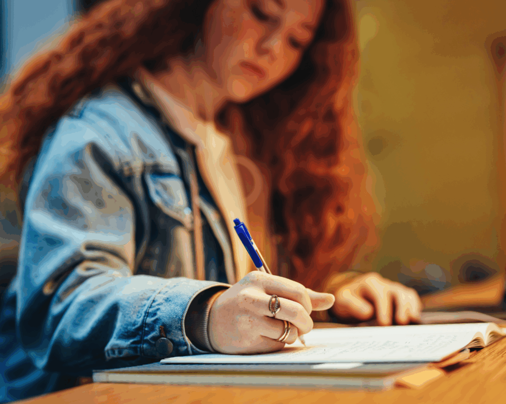 A warmly-lit photo of a college-age girl writing in a notebook at a desk in front of a laptop.
