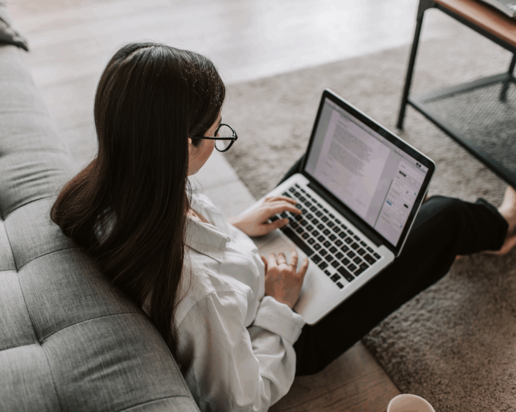 Over the shoulder photo of a student sitting on the floor of her home, with her laptop on her legs, typing in a document.
