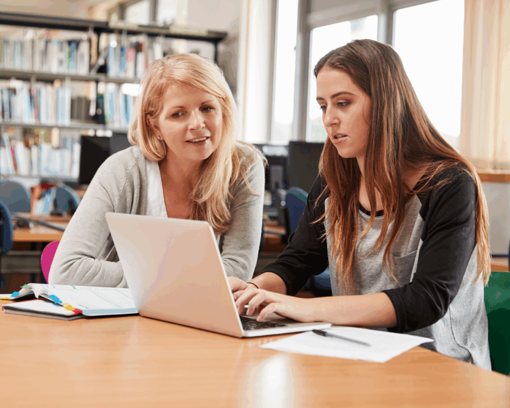 Bright photo of a college student and teacher seated together at a table, working together intently over a laptop.