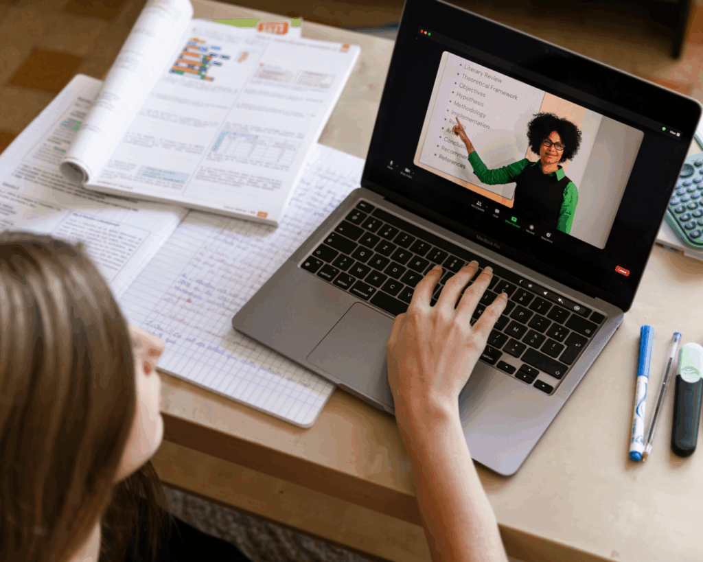 Over-the-should photo of a girl using a laptop on a tabletop, she's watching a lecture of a teacher gesturing at a whiteboard, and organized papers are spread around the computer.