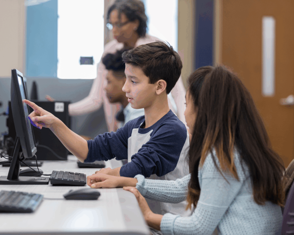 Photo of two middle school-aged children at a long computer lab desk, gesturing towards a shared computer monitor.