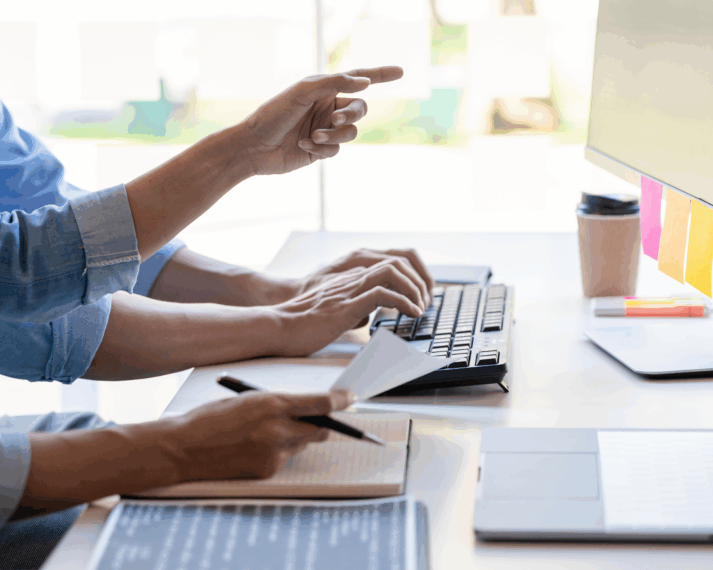Photo of two people's hands working together at a brightly lit desk. They type on a keyboard, hold a pen over paper, and gesture towards a computer monitor.