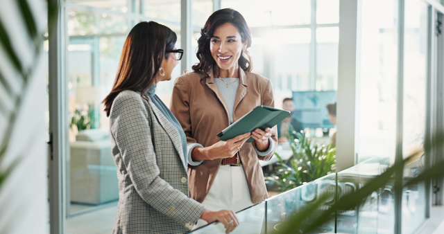 A brightly lit photo of two professionally-dressed women discussing paperwork near the clear railing of an office-space balcony.