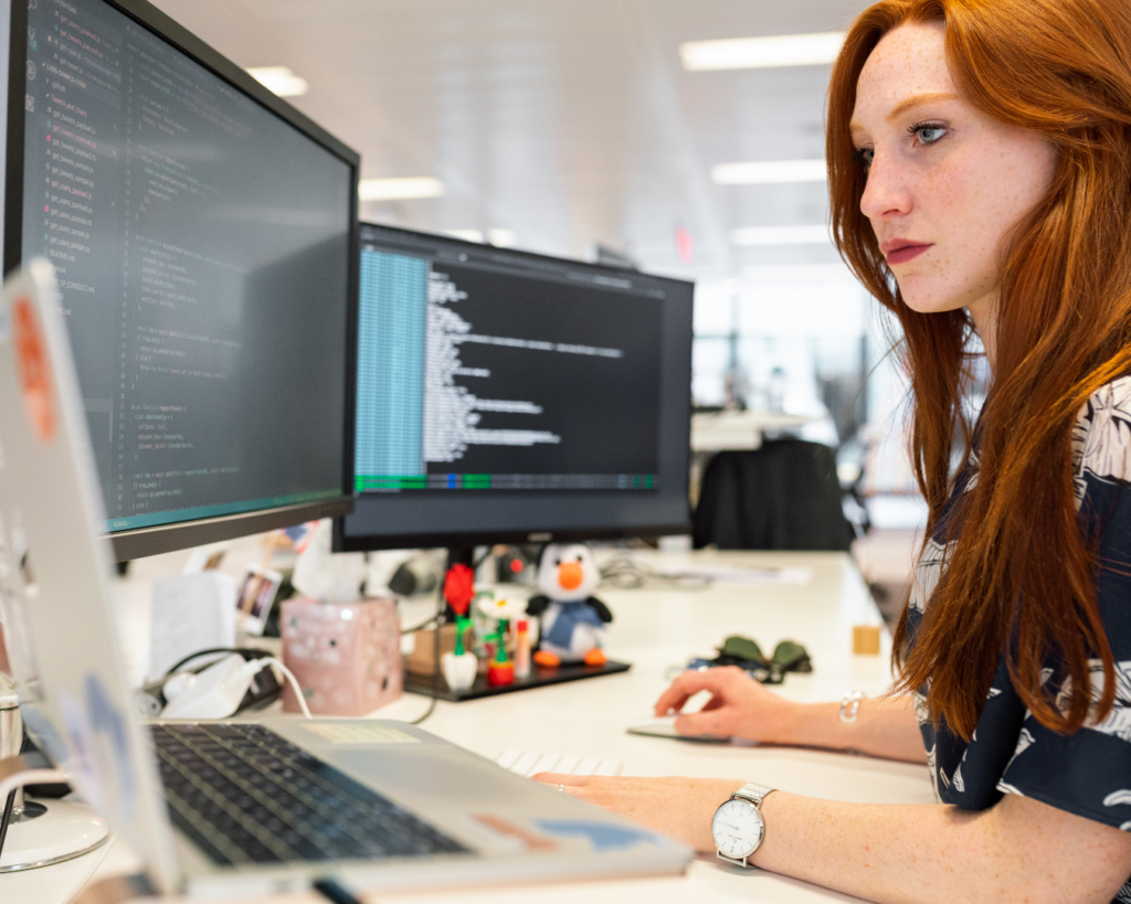 Photo of a focused women working at a colorfully cluttered desk with two computer monitors and an open laptop.