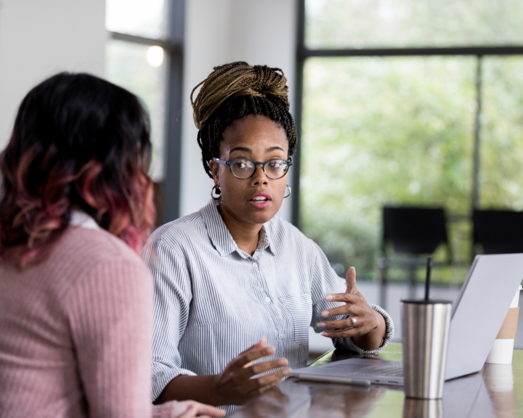 Photo of two professionally dressed women in a brightly lit office deep in discussion in front of a laptop.
