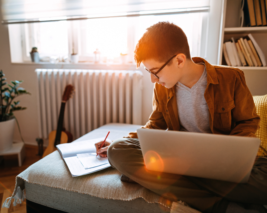 A warmly lit photo of a young man sitting on an ottoman with a laptop in his lap, leaning to the side to take notes in a notebook.