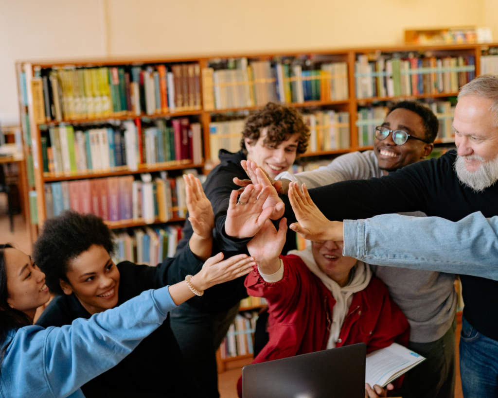 Photo of a diverse group of students in a library with their older professor sharing a group high-five.