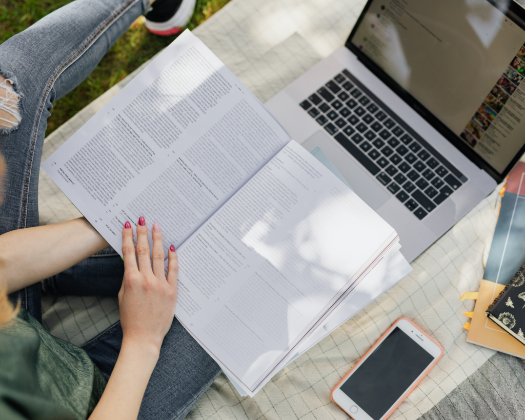 Birds-eye photo of a student seated on the ground with an open book and laptop in front of them, scanning the book page with their finger.