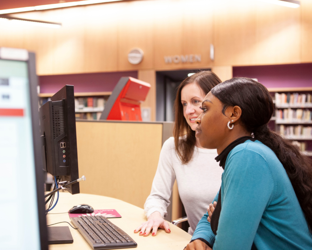 Photo of two women in a bright library sharing a desktop computer.