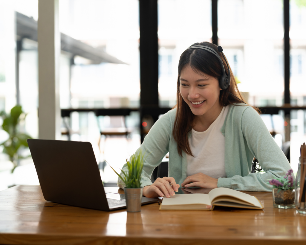 A photo of a smiling young college-age girl wearing headphones and smiling at her open laptop, pencil in hand.