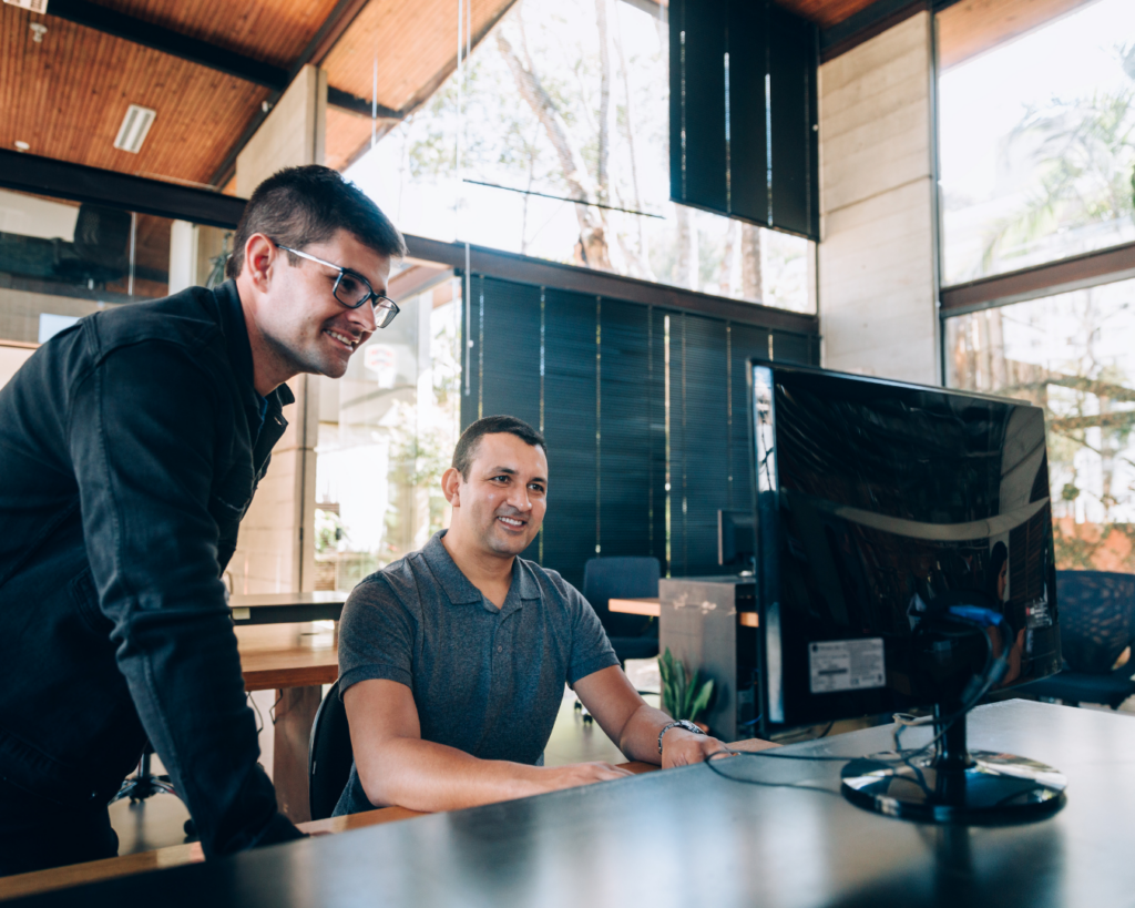 Bright photo of two men in front of a computer monitor. On is seated, the other stands over him, helping him work.