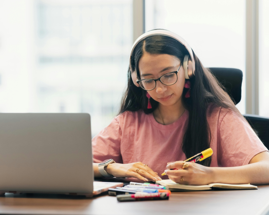 Photo of a young female college student studying at a table with an open laptop, she wears headphones and highlights text on a printout